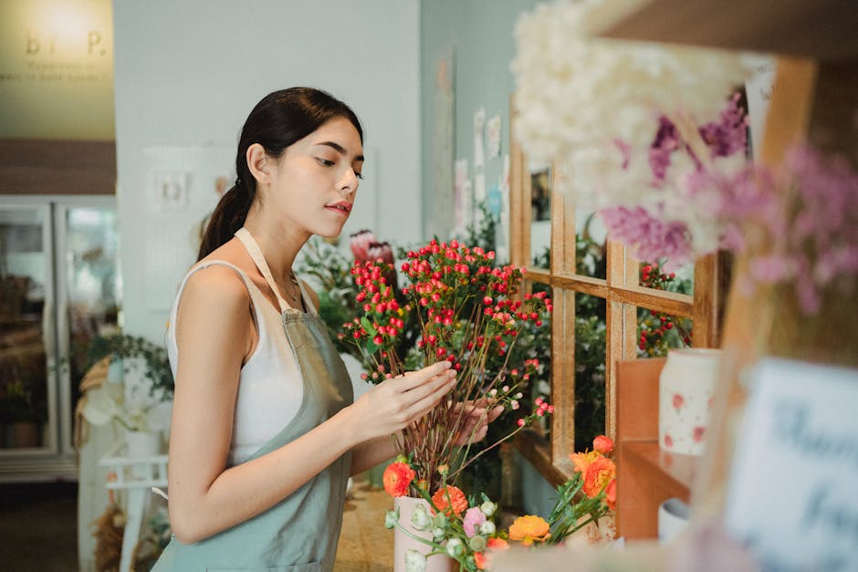 A young woman with dark hair tied back, wearing a light grey apron over a white sleeveless top, examines a bouquet of small, vibrant pink and red flowers with green stems inside a florist shop. She is