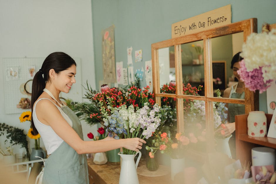 A young female florist with dark hair tied back, wearing a light green apron over a white top, is arranging a bouquet of fresh flowers on a wooden counter inside a florist shop. The bouquet includes r