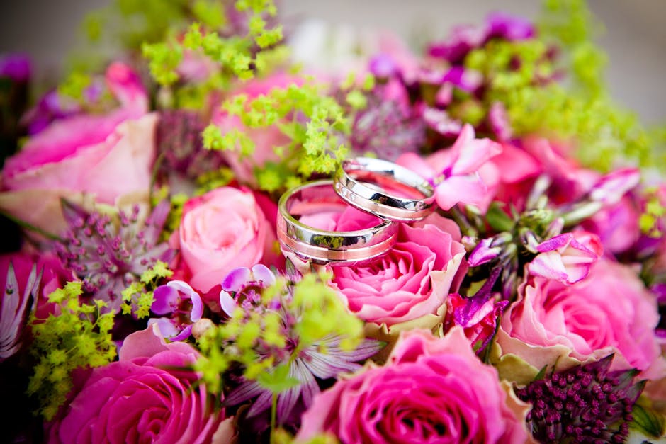 A close-up of two silver wedding bands resting atop a vibrant floral arrangement consisting of pink roses, purple orchids, and green foliage. The bouquet is lush and fresh, with the flowers arranged c