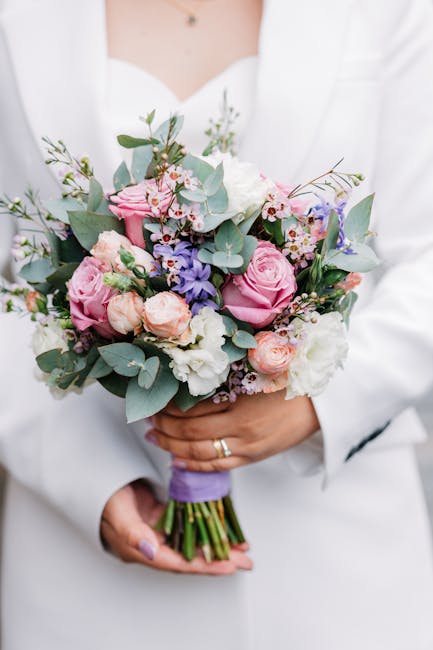 A person dressed in a white outfit holding a floral bouquet arrangement featuring pastel pink roses, white lisianthus, and light purple flowers complemented by green eucalyptus leaves. The bouquet is