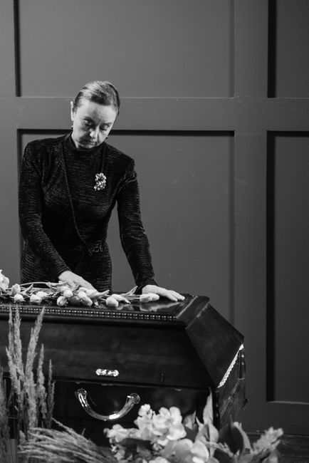 A woman dressed in black, with neatly styled hair and a floral brooch, is arranging a floral tribute on top of a closed casket during a funeral ceremony. The arrangement includes light-colored roses a
