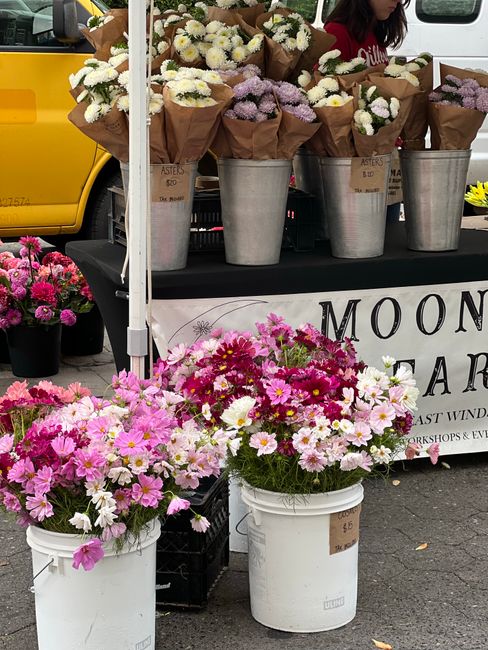 A display of fresh flower bouquets at Greenwich Market, arranged in large metallic buckets on a black table, with a variety of blooms including white chrysanthemums, pale purple flowers, and pink and 