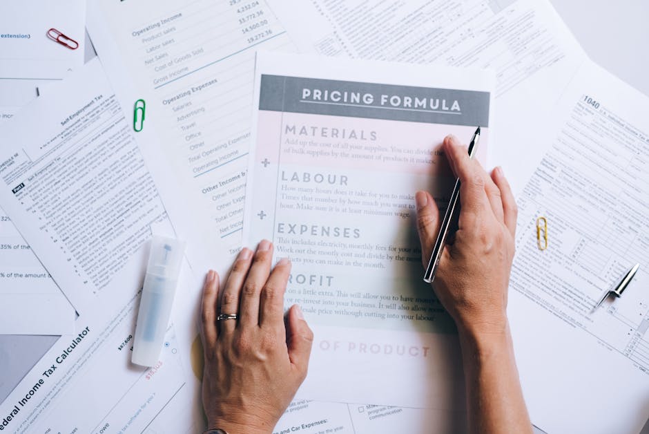 A close-up of a person's hands reviewing a printed pricing formula document amidst scattered financial papers, including tax calculators and expense spreadsheets, on a white desk. The person's right h