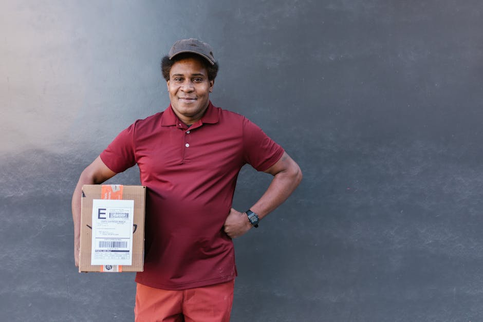 A delivery person with dark skin, short curly hair, and a friendly smile, dressed in a maroon polo shirt and matching shorts, holding a cardboard parcel with a delivery label in their left hand. The p